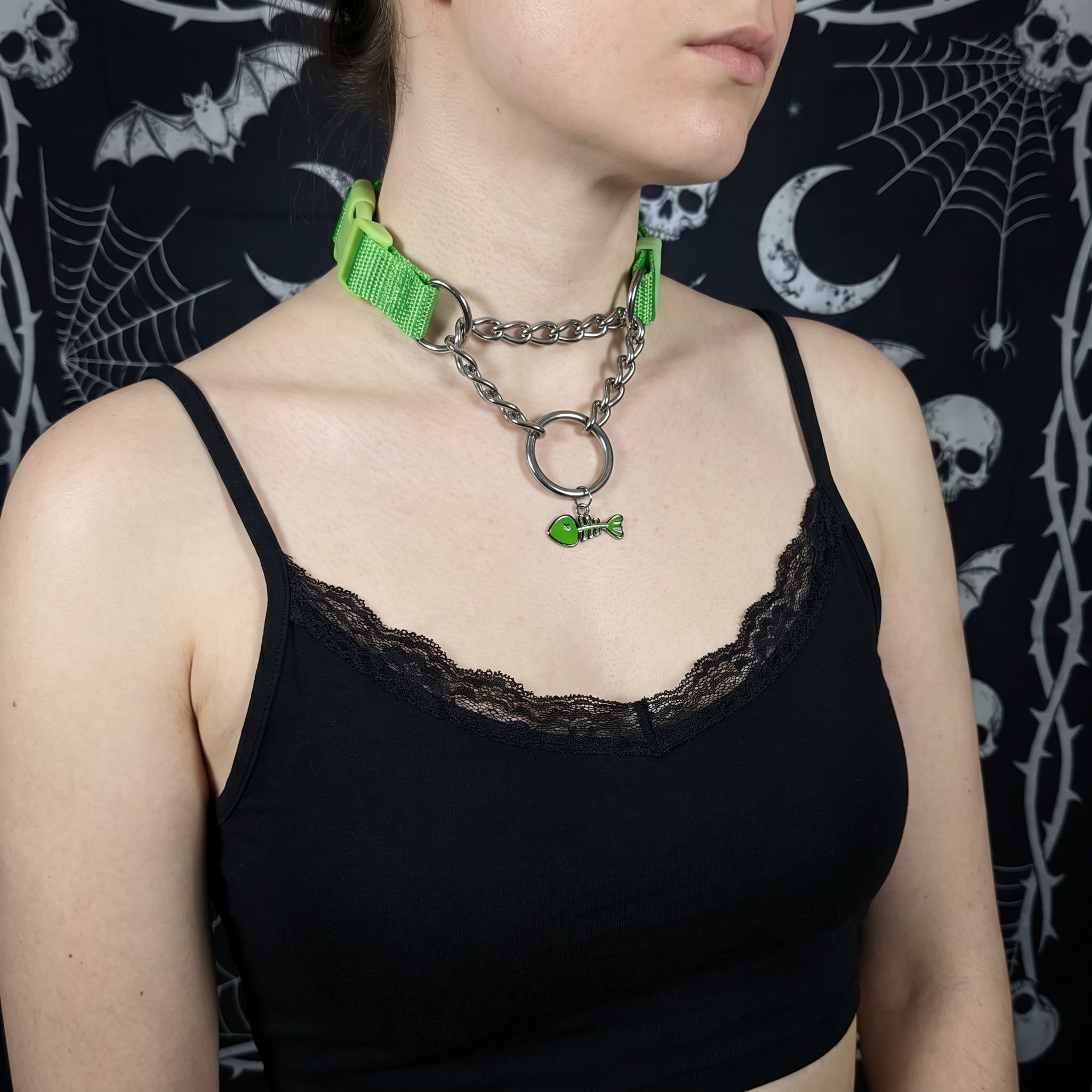 Person wearing a black tank top with a green collar and chain against a dark background with spider webs and bats.