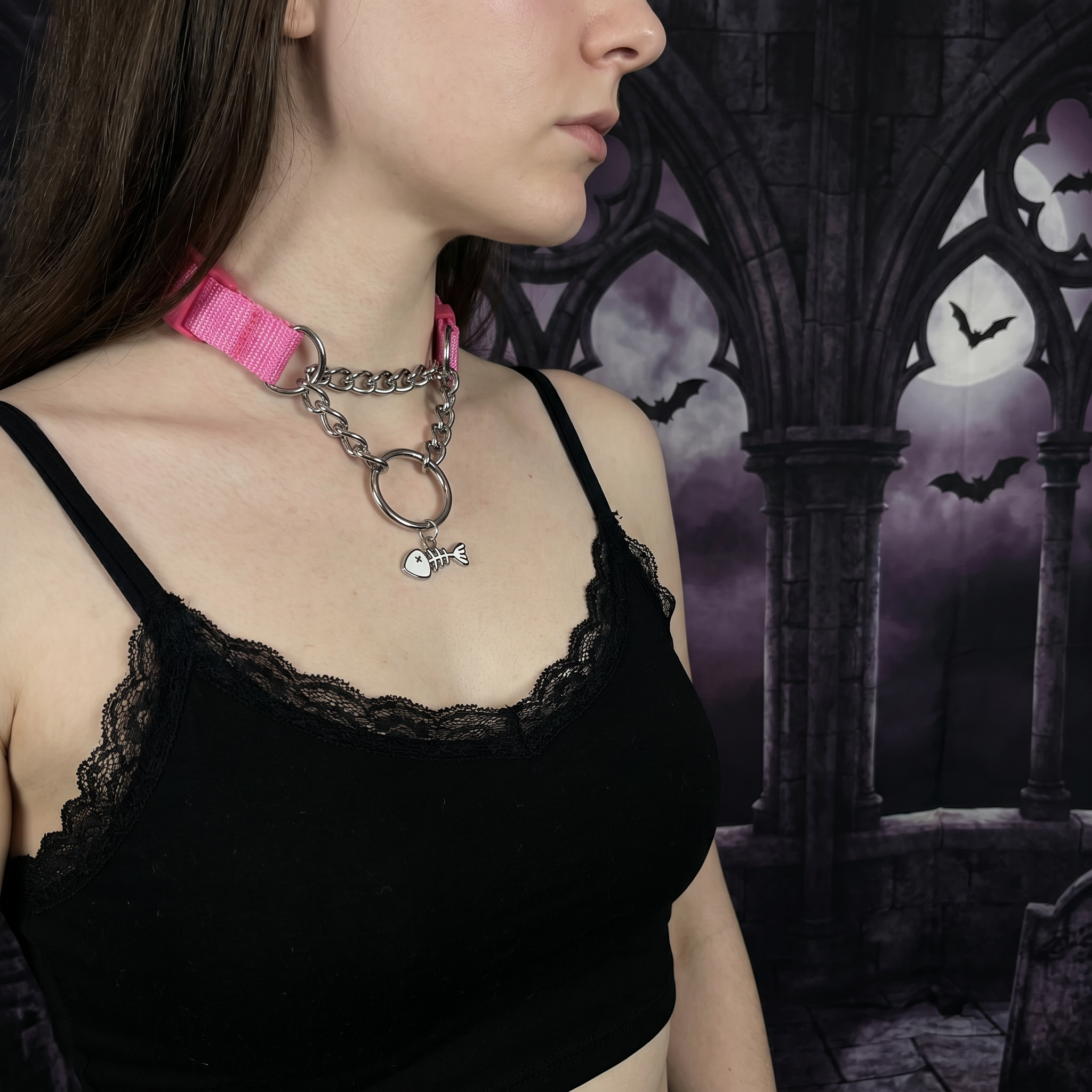 Person wearing a black lace top with a pink choker and chain necklace against a gothic-style background.
