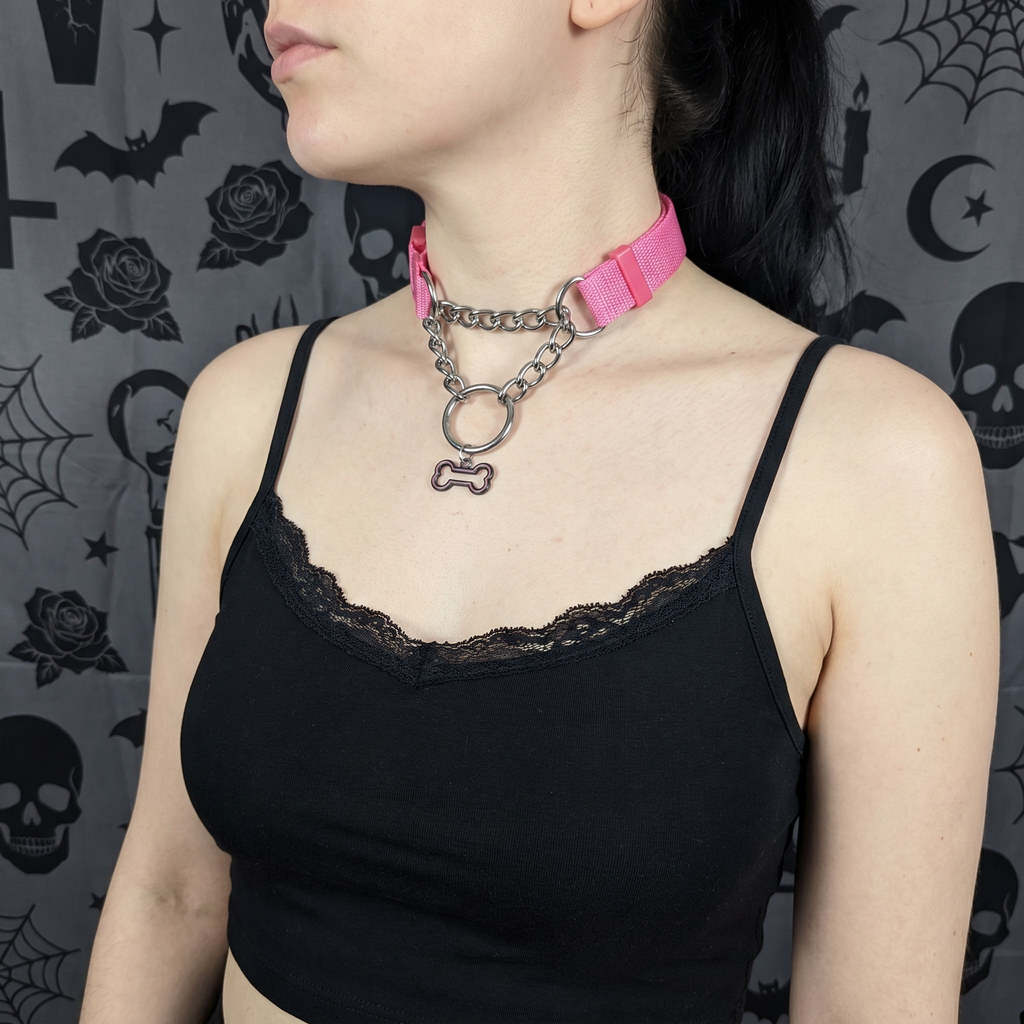 Person wearing a pink choker with a chain and bone pendant against a dark, patterned background.