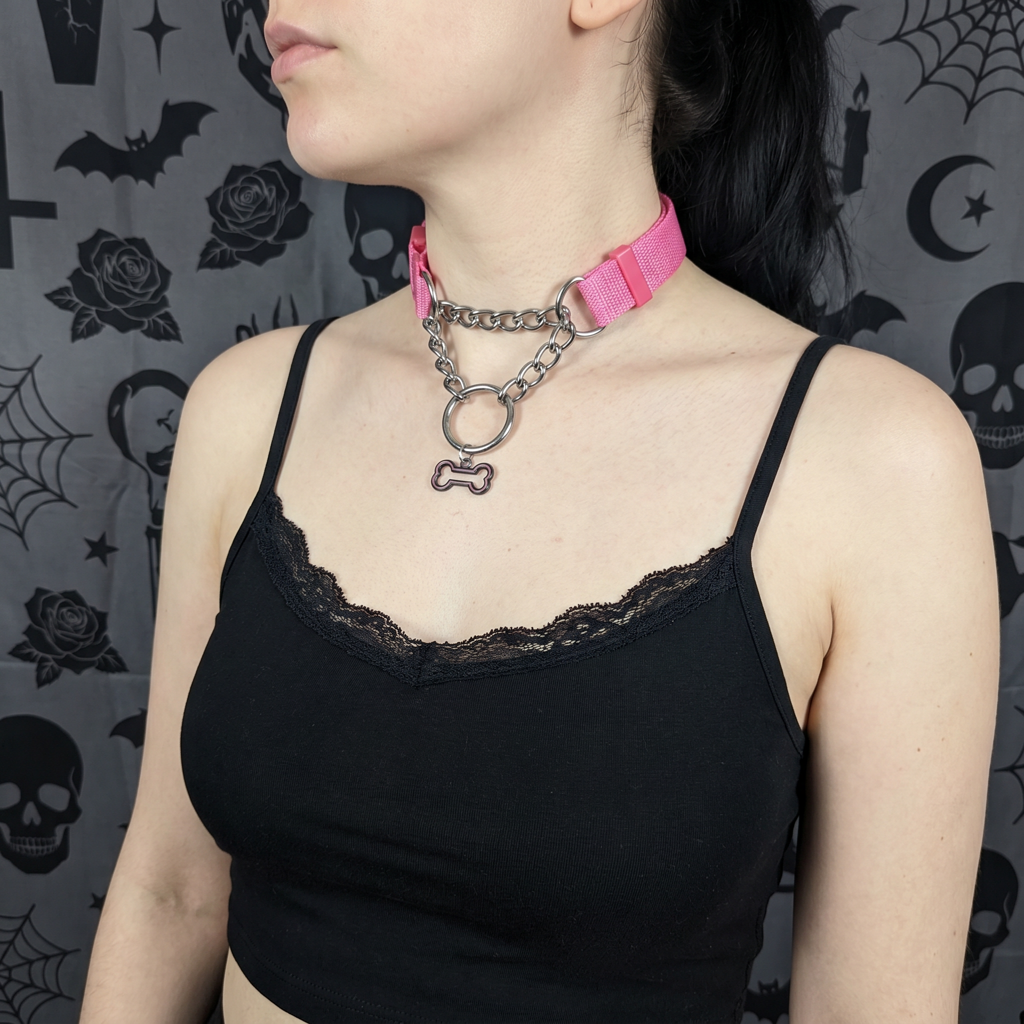 Person wearing a pink choker with a chain and bone pendant against a dark, patterned background.